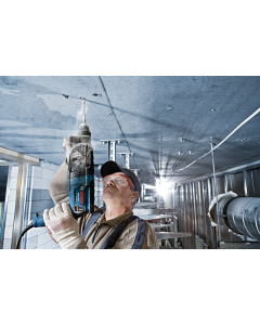 A man wearing a baseball cap and safety glasses using a Bosch GBH 2-24D Rotary Hammer SDS Drill to drill into a ceiling. 