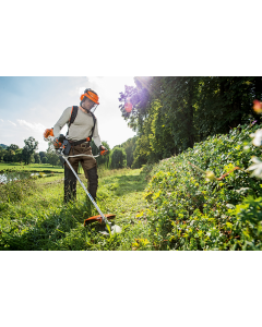 A man using a Stihl FS 131 Petrol Brush Cutter on a large area of overgrown grass and shrubbery. The man is wearing protective gloves