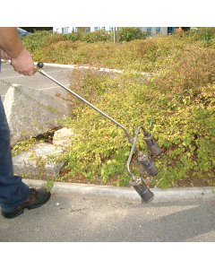 Man in navy trousers and safety footwear using a three-head flame gun on a section of shrubbery adjacent to a car park. 