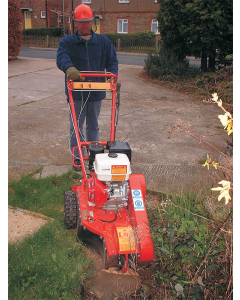 Man wearing protective gloves, red safety helmet and visor using a red petrol stump grinder a small patch of grass with a driveway and residential properties in the background.